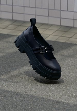 Černá masivní bota typu Mary Jane s dvojitými stříbrnými přezkami na texturovaném povrchu poblíž obložené zdi.