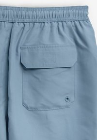 Pantaloni scurți de culoare albastră deschisă cu talie elastică, având o buzunar la spate cu clapă și un mic detaliu cu un ochișor aproape de marginea buzunarului.