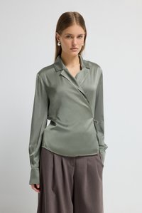 Bluse aus Satin in gedämpftem Grün mit langen Ärmeln, einem Reverskragen und seitlicher Schleifendetail. Kombiniert mit locker sitzenden grauen Hosen.