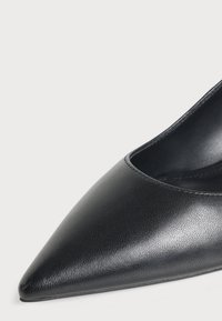 Gros plan d'une chaussure en cuir noire à bout pointu, avec une texture lisse et peu de coutures sur un fond clair.