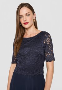 Abito blu navy con top in pizzo a maniche corte, motivi floreali testurizzati e un tessuto inferiore liscio che crea un effetto a strati.
