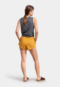 Grijze mouwloze top, gele shorts met een gladde textuur en zwarte sandalen. De shorts hebben een ontspannen pasvorm en zijzakken.