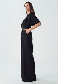 Ženska stoji v črnem širokem kombinezonu z kratkimi širokimi rokavi in se postavlja z eno roko v žepu pred enotno ozadje.