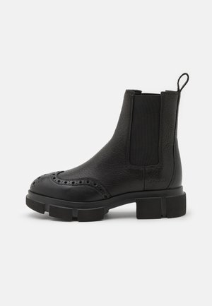 Stivale alla caviglia in pelle nera con pannelli elastici sui lati, finitura testurizzata, dettagli brogue, suola in gomma spessa e un manico sul retro.