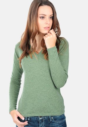 Pull vert en V en tissu doux et texturé avec des manches longues; associé à un jean en denim foncé, mettant en valeur une silhouette ajustée.