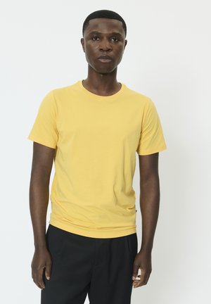 Jeune homme debout, portant un t-shirt jaune uni à manches courtes et un pantalon foncé sur un fond blanc.