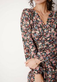 Kwiecista sukienka z dekoltem w serek, długimi bufkowatymi rękawami. Na ciemnozielonym tle znajdują się różowe, niebieskie i białe kwiaty. Do kompletu dołączona jest złota bransoletka.