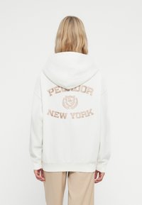 Bijela hoodie s izvezenim tekstom "PEQUODOR NEW YORK" na leđima. Mekana, teksturirana tkanina s prostranom kapuljačom i rebrastim manžetama.