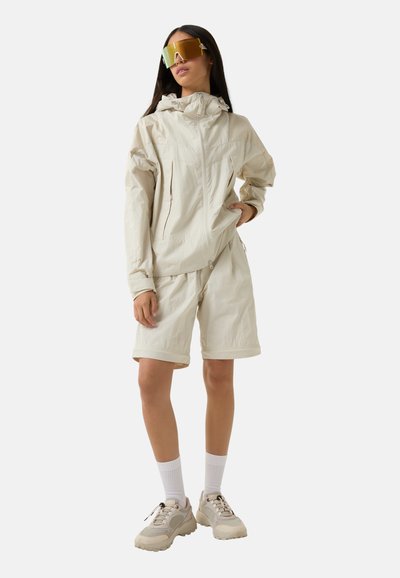 Beige letvægtsjakke med hætte, lynlåslukninger og lommer, parret med matchende shorts. Modellen bærer oversized guld solbriller og sneakers.