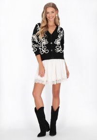 Čierny cardigan s bielymi vyšitými slučkami, s výstrihom do V a gombíkmi, v kombinácii s bielymi šatami s čipkovaným lemovaním a čiernymi semišovými členkovými botami.