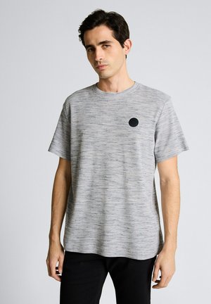 Mężczyzna w jasnoszarej teksturowanej koszulce z krótkim rękawem z małym czarnym okrągłym logo na lewej piersi, stojący na jednolitym tle.