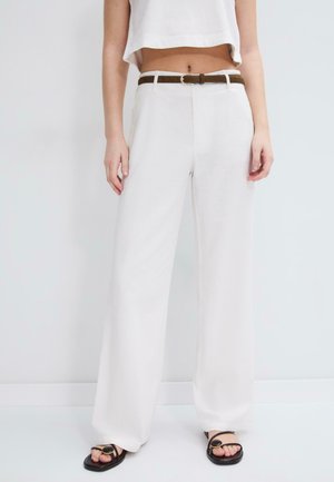 Osoba nosiaca široké biele nohavice s vysokým pásom, hnedý opasok, krátky biely top a čierne sandále s otvorenou špičkou, stojaca proti jednoliatemu pozadiu.