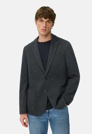 Donkerblauwe blazer van een gestructureerde stof, met een gelapte kraag, twee voorkruiszakken, een enkele knoopsluiting, gecombineerd met een zwart shirt en blauwe jeans.