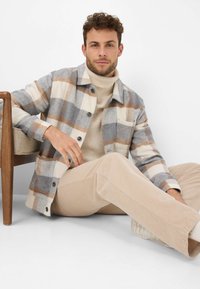 Giacca a camicia a quadri in grigio, beige e marrone; chiusura frontale con bottoni, tasca singola, indossata sopra un dolcevita beige e abbinata a pantaloni in corduroy beige.