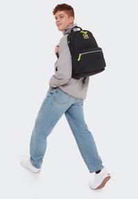 Zaino nero con dettagli verdi, cerniere e tessuto testurizzato, indossato su una spalla; modello con giacca grigia chiara e jeans blu.