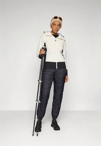 Weiße isolierte Jacke mit schwarzen Akzenten, kombiniert mit schwarzen gepolsterten Hosen. Hält Trekkingstöcke, trägt einen beigen Hut und eine Sonnenbrille.