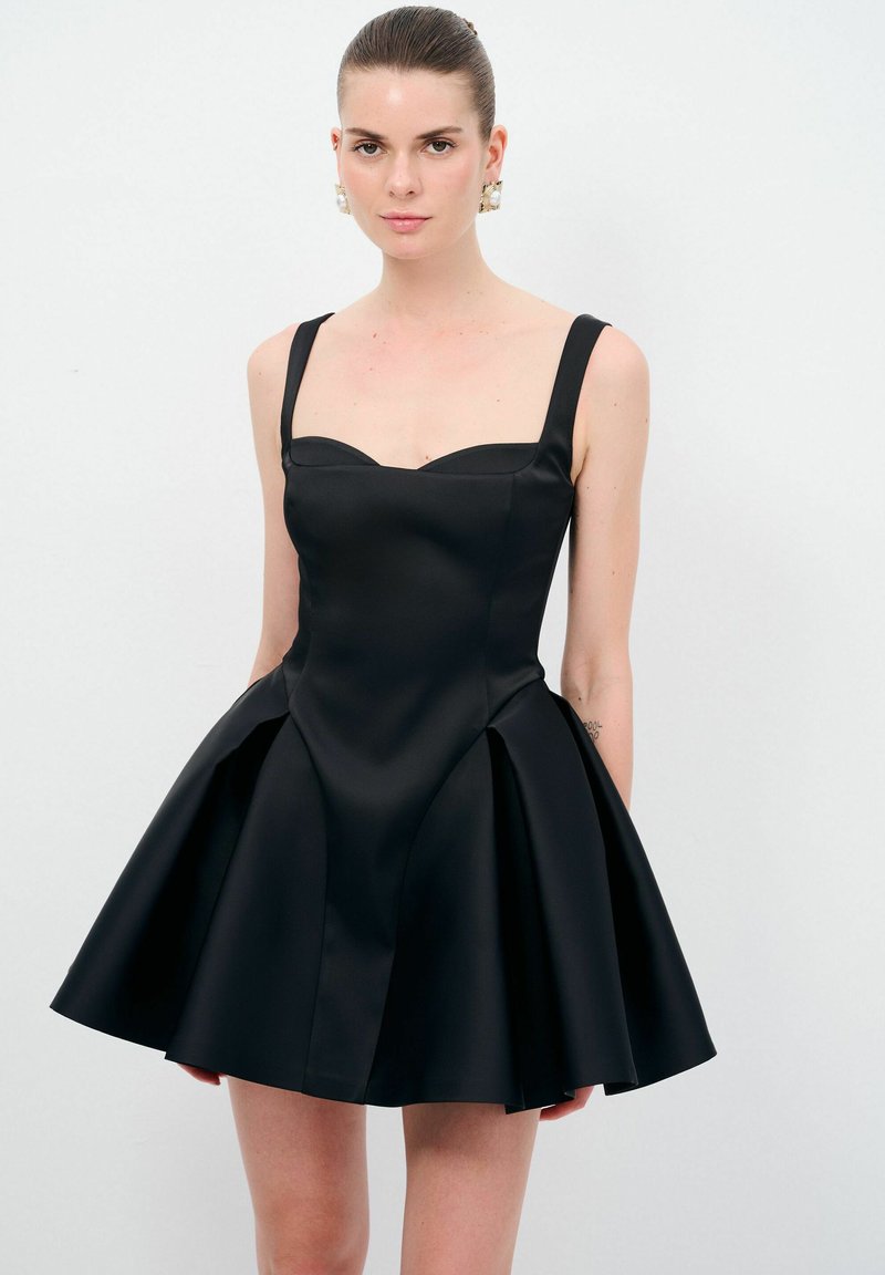 Robe en satin noir avec un corsage ajusté et une jupe évasée. Présente de larges bretelles, un décolleté en cœur et des détails de coutures visibles.