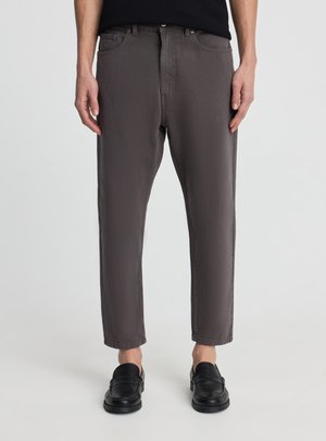 Mężczyzna ubrany w krótkie ciemnoszare spodnie i czarne skórzane mokasyny, stojący na tle jasnego, jednolitego tła.