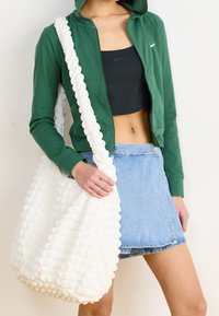 Vit texturerad väska med en vågig rem, bärs över en grön zip-hoodie och svart crop top, kombinerad med en blå jeanskjol.