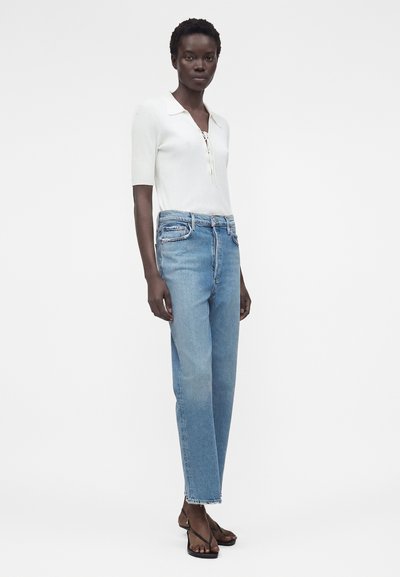 Γυναίκα που φοράει ένα λευκό ριμπ πλεκτό τοπ με κοντά μανίκια και κορδόνια μπροστά, ανοιχτό μπλε ψηλόμεσο τζιν με ίσια γραμμή και μαύρα σανδάλια με λουράκια, στέκεται κοιτώντας προς τα εμπρός.