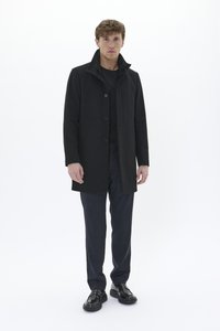 Manteau noir avec un col haut, doté d'une fermeture à boutons et de poches latérales. Porté par-dessus un haut noir et un pantalon à motifs sombres. Des chaussures noires complètent le look.