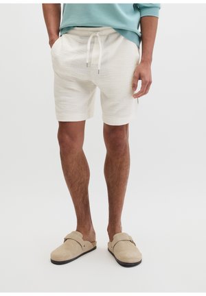 Mann trägt cremefarbene, strukturierte Shorts mit Kordelzug, hellblauen Pullover und beige Slipper vor einfachem Hintergrund.