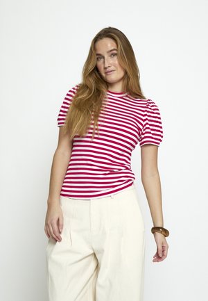 Junge Frau mit langen blonden Haaren, die ein rot-weiß gestreiftes Hemd und cremefarbene, weit geschnittene Hosen trägt, posiert vor einem weißen Hintergrund.