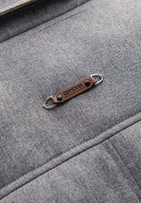 Tela gris con una textura suave que presenta una etiqueta de cuero marrón con acentos metálicos mostrando la palabra "ombre" en negro.