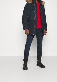 Námořnická parka s kapucí lemovanou kožešinou, několika předními kapsami, zapínáním na knoflíky, v kombinaci s červenou košilí, tmavými džínami a černými botami.