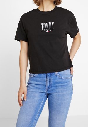 Kobieta ubrana w czarną koszulkę Tommy Jeans i jasnoniebieskie, wysokie jeansy, stoi z jedną ręką w kieszeni, na białym tle.