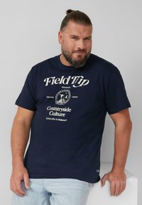 Mężczyzna z brodą i związanymi włosami, ubrany w granatowy t-shirt "Field Trip Countryside Culture" oraz jasne dżinsy, oparty o białą powierzchnię.