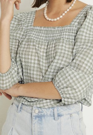 Blusa a cuadros verdes y blancos con mangas abullonadas y detalles fruncidos en el escote, combinada con vaqueros de mezclilla de lavado claro. Perlas adornan el escote.