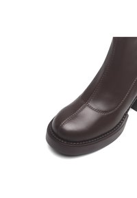 Primer plano de una bota de cuero marrón oscuro que muestra la punta redondeada, costuras detalladas, suela gruesa y tacón ancho sobre un fondo blanco.