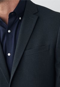 Blazer azul marino con superficie texturizada, un bolsillo en el pecho y solapas con muesca, combinado con una camisa azul marino ajustada abotonada.