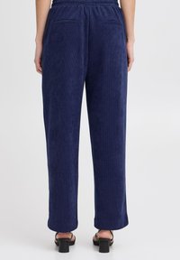 Pantalons en velours côtelé bleu marine avec une taille élastique, deux poches arrière et une coupe droite, associés à des sandales à talons blocs.