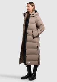 Long, beige puffer coat with horizontal stitching, a hood, zippered pockets, and black accents; paired with black knee-high boots.