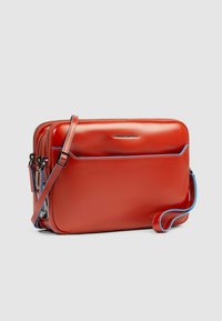 Rode leren crossbodytas met een glanzende afwerking, voorzien van twee met rits afsluitbare vakken, blauwe accenten en een afneembare riem.