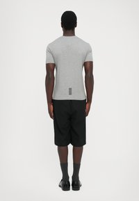 Mann med en tettsittende grå kortermet skjorte med svarte vertikale striper nær kanten, svarte kne-lange shorts, mørke sokker og sorte sko, som står med ryggen til.