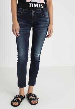 Jeans de mezclilla azul ajustados con detalles desgastados, cinco bolsillos y cierre de botón, combinados con sandalias negras con hebillas.