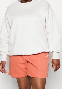 Ljusbeige sweatshirt med rund halsringning och vitt Nike-swoosh-logotyp; ihop med korallfärgade shorts med sidfickor och logotypaccent.