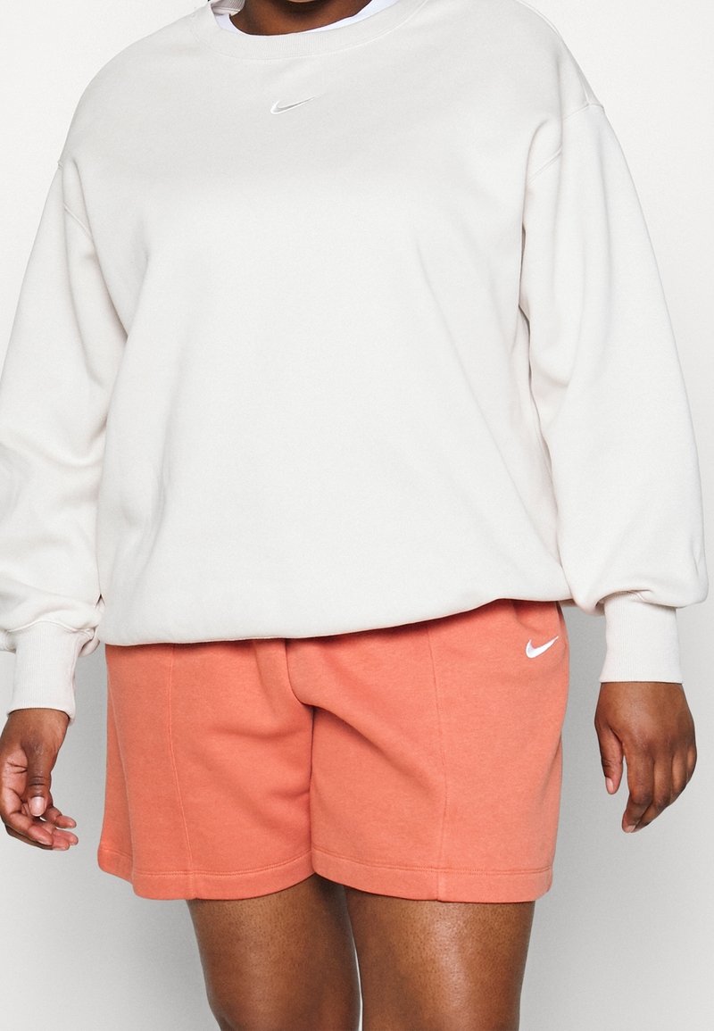 Ljusbeige sweatshirt med rund halsringning och vitt Nike-swoosh-logotyp; ihop med korallfärgade shorts med sidfickor och logotypaccent.