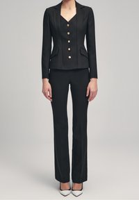 Blazer noir ajusté avec des boutons dorés, doté d'un décolleté en V et de manches longues, associé à un pantalon noir ajusté et des talons pointus.
