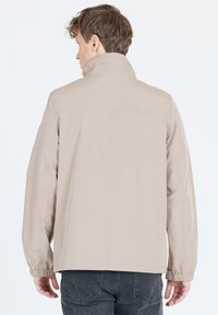 Beige jacka med hög krage, elastiska muddar och sömmar för struktur. Tyget verkar glatt och hållbart. Bärs över mörka jeans.