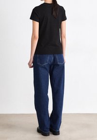 Frau von hinten, trägt ein schwarzes kurzärmliges Shirt, blaue gerade Jeans und schwarze Schuhe, steht auf einem hellen Boden vor einer weißen Wand.