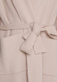 Cardigan en tricot côtelé couleur crème, noué à la taille avec une ceinture assortie, présentant deux poches avant.