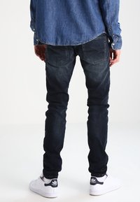 Jeans de denim oscuros de corte ajustado y acabado desgastado, con dos bolsillos traseros y un aspecto usado. Combinados con zapatillas blancas.