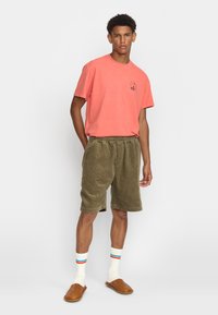 Koraalkleurig T-shirt met korte mouwen, zakdetail. Olijfgroene ribfluwelen shorts. Witte sokken met gekleurde strepen. Bruine instappers.