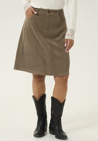 Jupe A-line en corduroy marron avec poches avant, passants de ceinture et texture légèrement côtelée. Associée à des bottes de cowboy noires à zip.