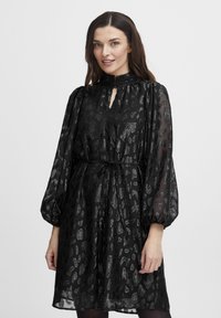 Rochie neagră dintr-un material transparent, texturat, cu un imprimeu animalier subtil. Gât înalt, detaliu cu tăietură în formă de cheie și mâneci bufante, cu un cordon la talie.