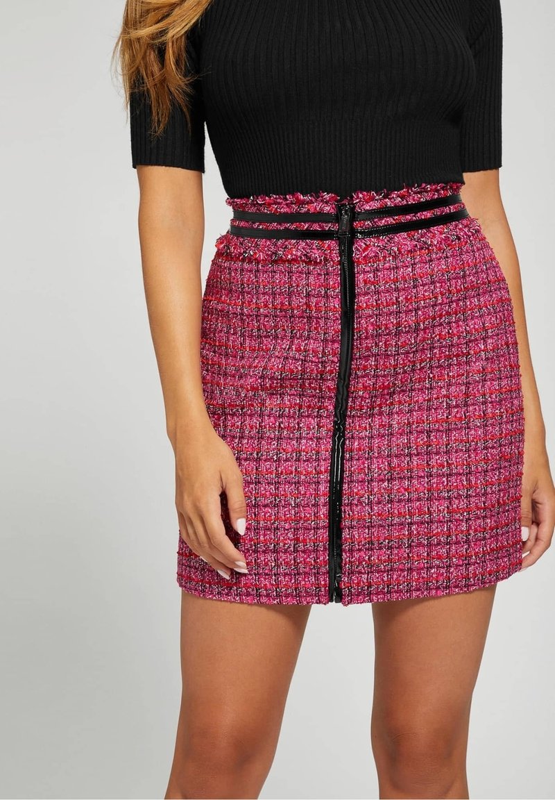 Femme portant une mini-jupe texturée rose avec une fermeture éclair verticale noire et une ceinture noire à la taille, associée à un haut côtelé noir à manches courtes.